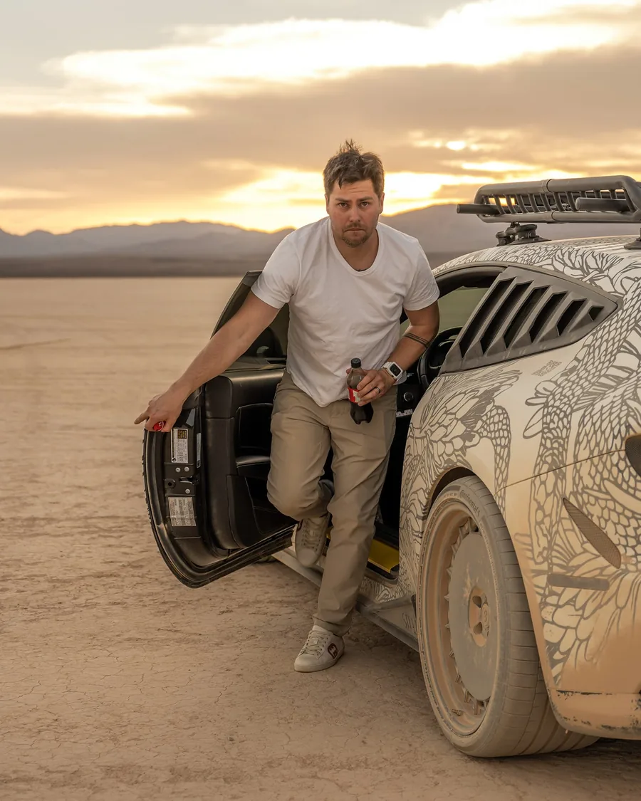 Trevin Hermosillo stepping out of a rally car on a dry lake bed at sunset.